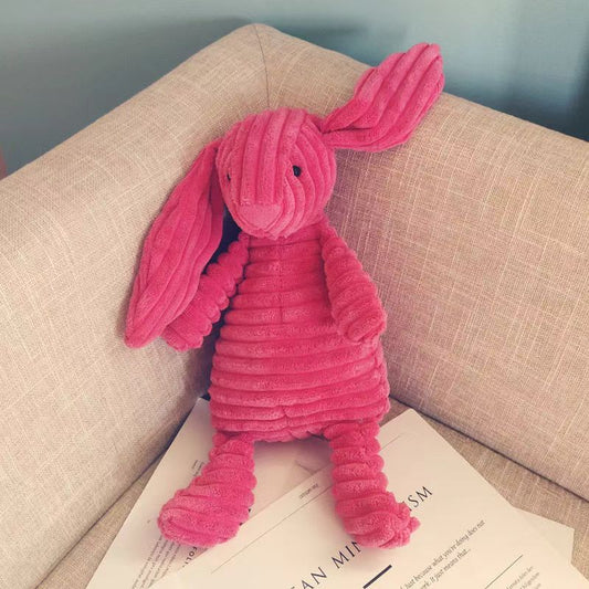 Pink corduroy plush bunny stuffed animal toy with long floppy ears sitting on a beige sofa, soft cuddly rabbit doll for kids and nursery decor.